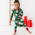 Child wearing a green Christmas-themed pajama set with red cuffs and trim, standing in front of a white wall with a wooden ladder and red gift boxes.
