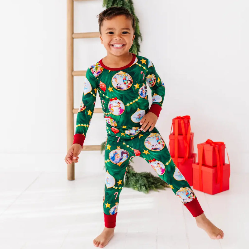 Child wearing a green Christmas-themed pajama set with red cuffs and trim, standing in front of a white wall with a wooden ladder and red gift boxes.
