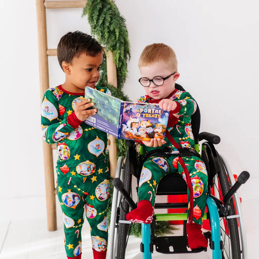 Two children in matching green pajamas with festive designs are reading books together, one in a wheelchair.