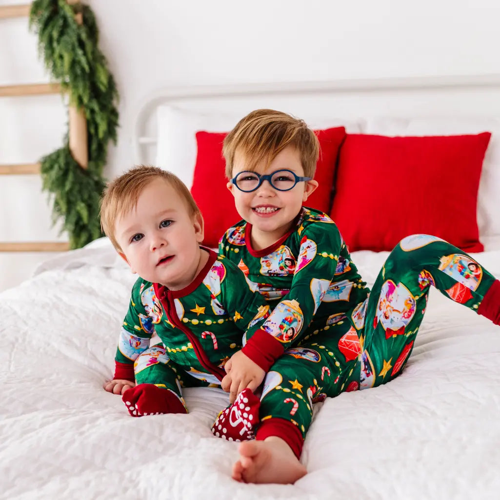 Two children in festive pajamas sitting on a bed with red pillows and Christmas decorations.