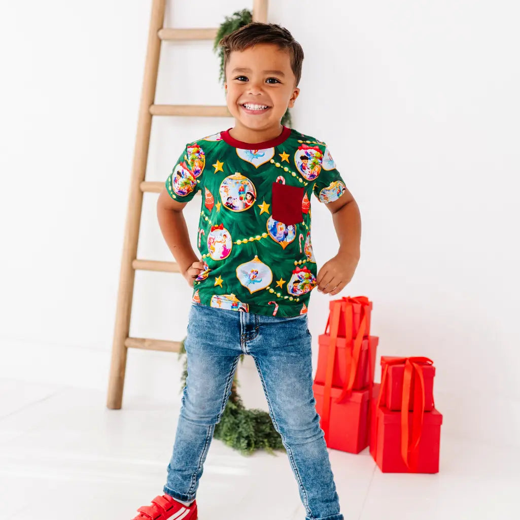 Child wearing a festive green shirt with colorful designs, standing next to a wooden ladder and red gift bags.