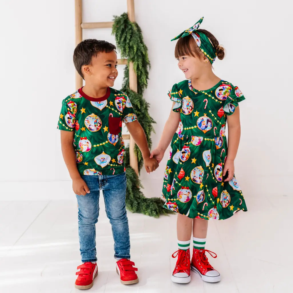 Two children holding hands, wearing matching festive outfits with a green background.