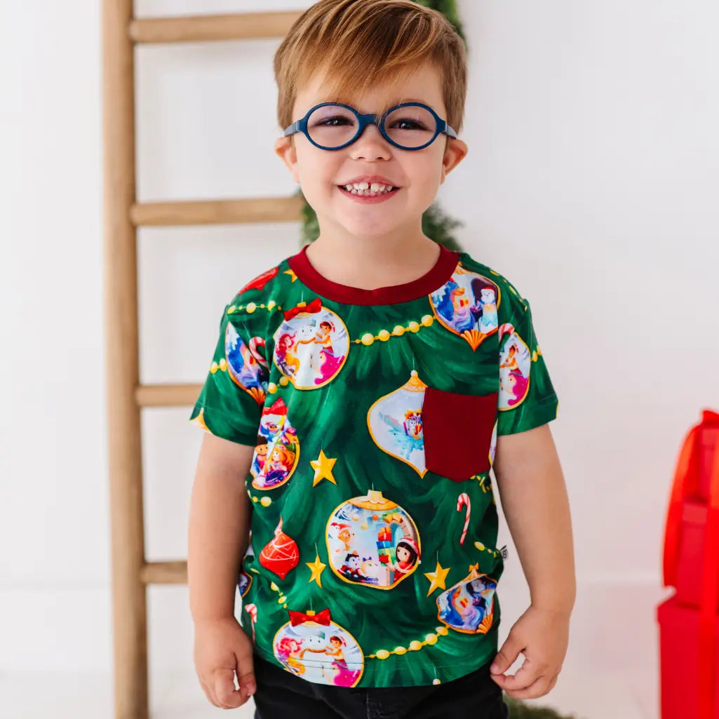 Child wearing a festive green shirt with colorful designs, standing against a white wall.