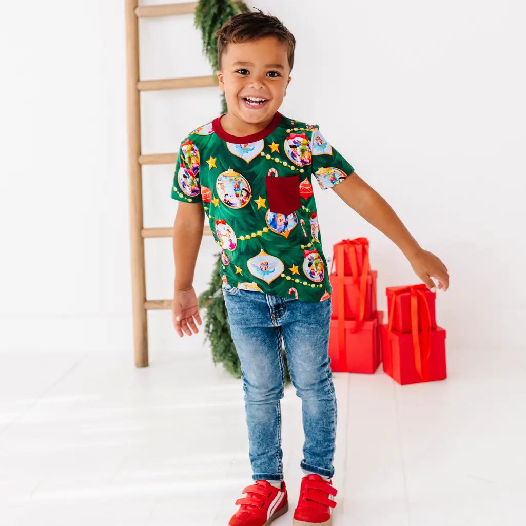 Child wearing a colorful Christmas-themed shirt with red shoes and blue jeans, standing next to a wooden ladder and Christmas decorations.