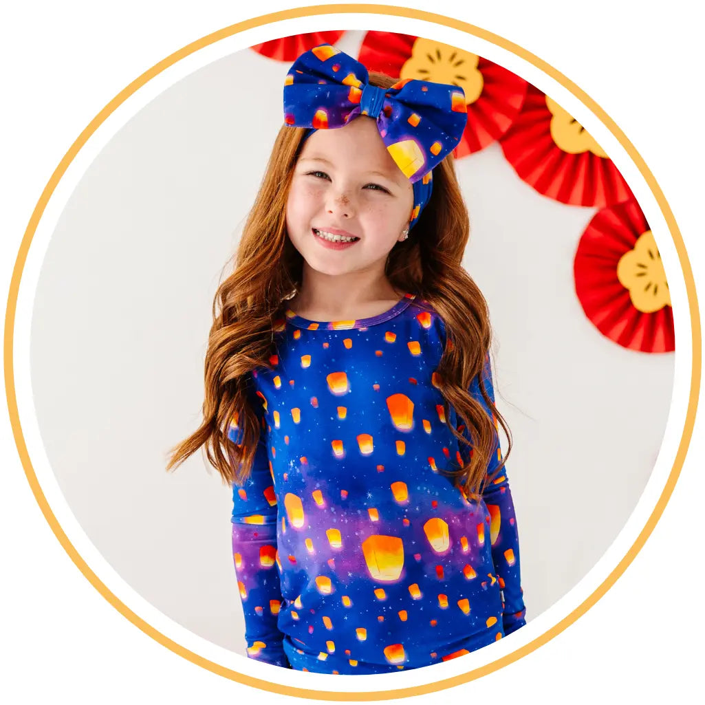 Young girl wearing a colorful dress with lantern pattern and headband, standing against a white background.