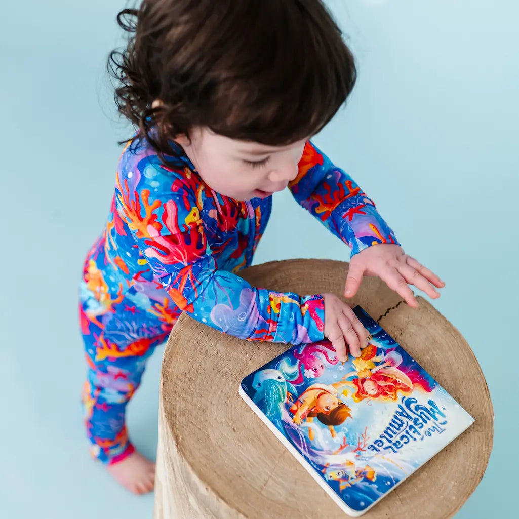Child in colorful outfit interacting with a book on a stool against a light blue background.