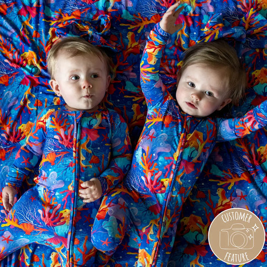 Two infants lying on a surface with a colorful coral-patterned pajama onesie, with the brand's watermark in the bottom right corner.