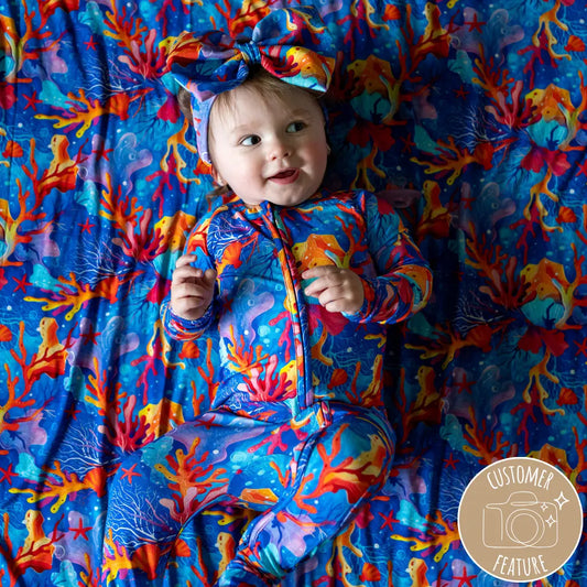 A baby wearing a colorful floral outfit with a bow on the head, lying on a matching floral blanket.