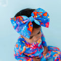 A baby wearing a colorful bow headband with a Reef-inspired pattern in blues, oranges, and purples.