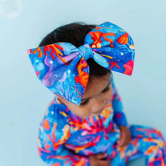 A baby wearing a colorful bow headband with a Reef-inspired pattern in blues, oranges, and purples.