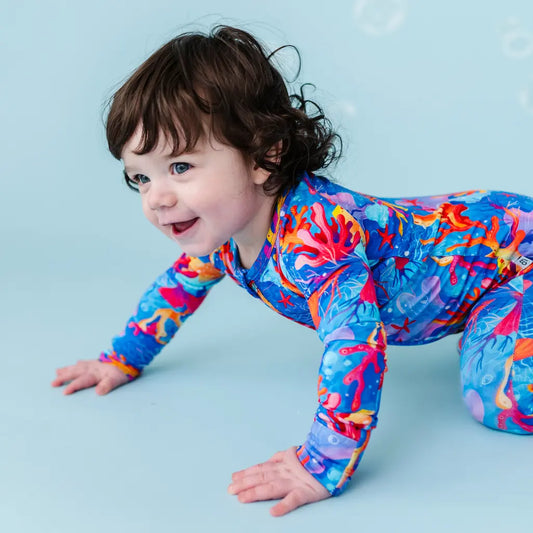 A baby wearing a colorful one-piece pajama with a pattern of vibrant corals and sea life in shades of blue, orange, and purple.