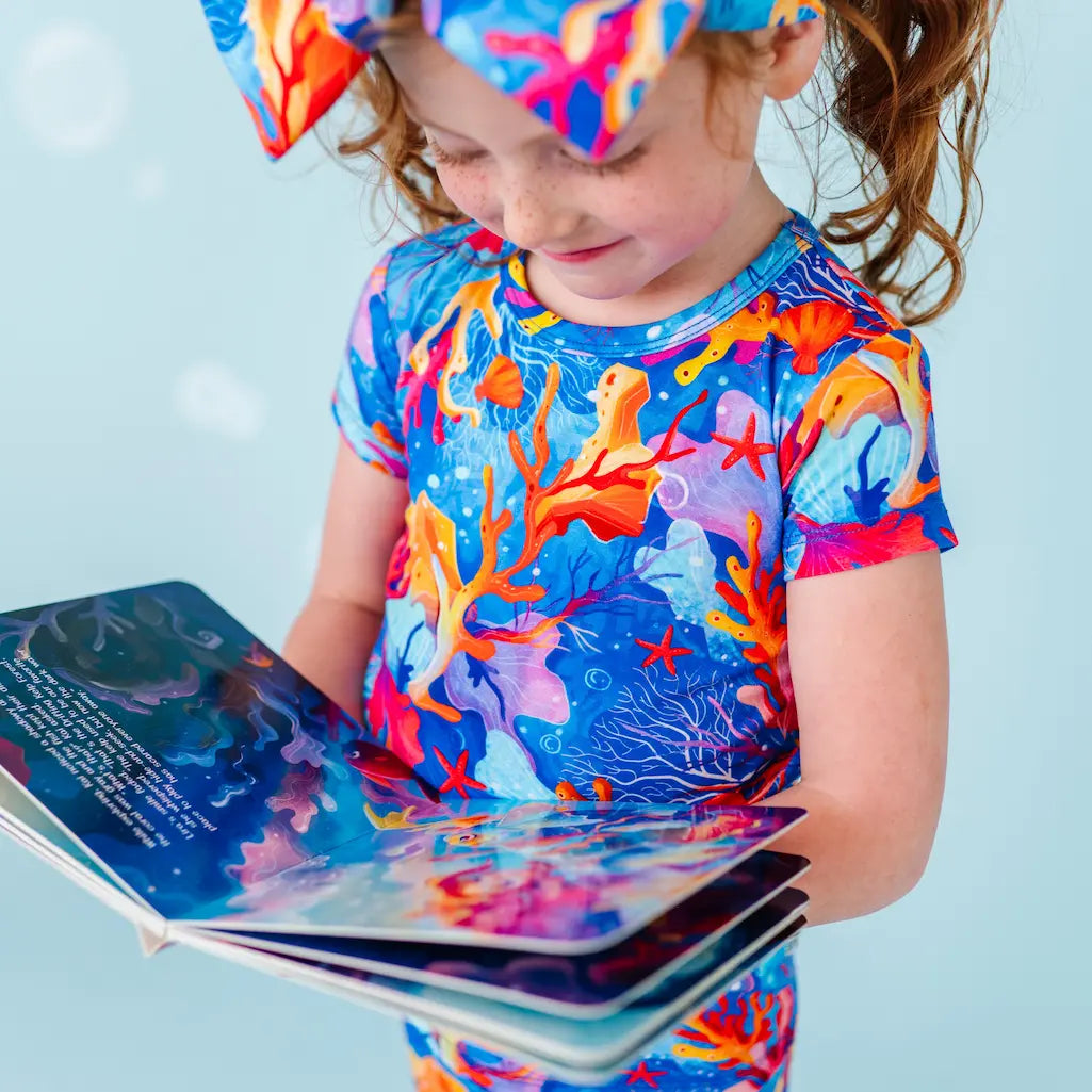 Child wearing a colorful coral-themed shirt and cap, holding a book.