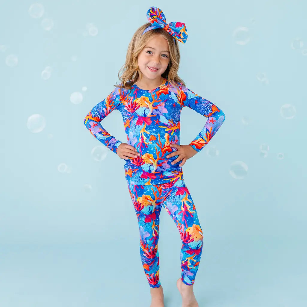 A child models a vibrant multicolored pajama set with a coral and sea creature pattern, consisting of a long-sleeve top and matching pants, accessorized with a hair tie.