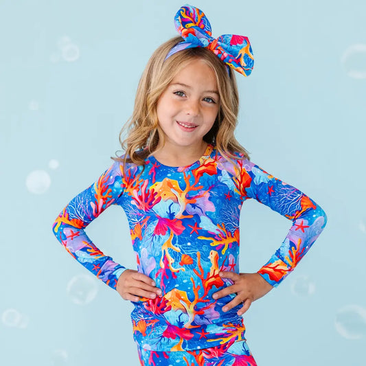 Child wearing a colorful coral-themed long-sleeve shirt and headband on a light blue background.