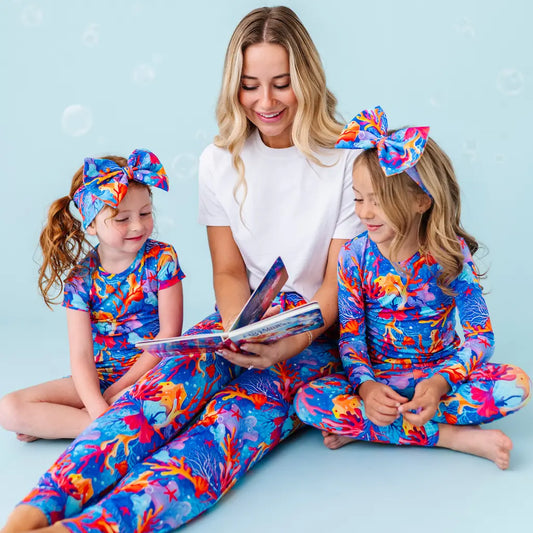 Woman and two children in colorful outfits reading a book against a light blue background.