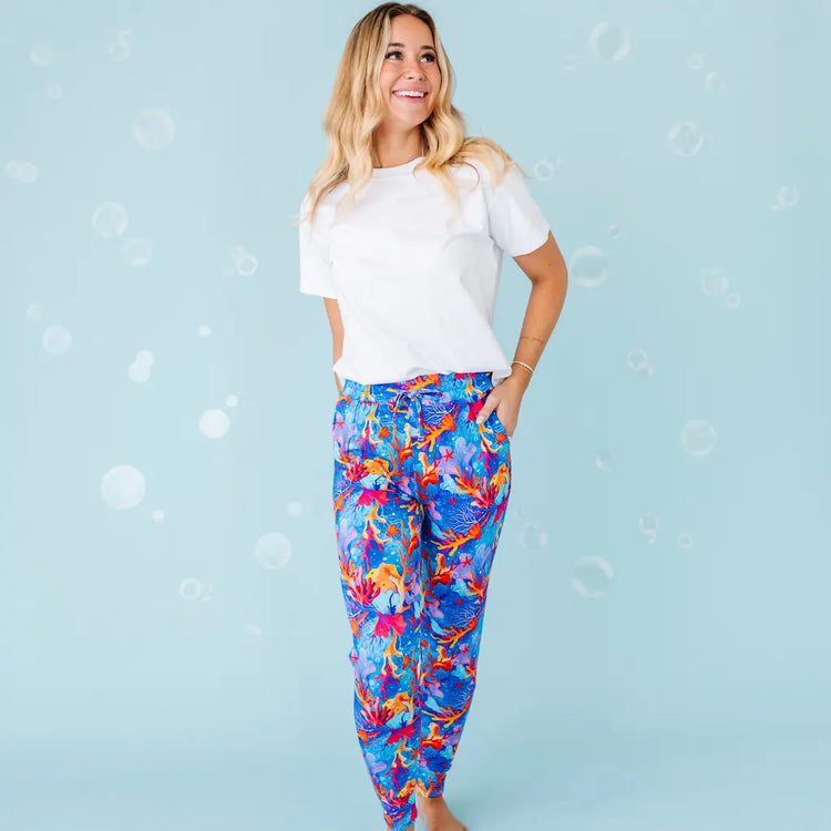 A woman standing and smiling, wearing white shirt and multicolored pajama pants with a coral reef pattern.