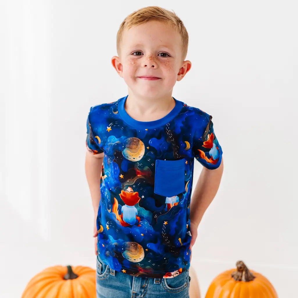 Child wearing a blue t-shirt with space-themed design, standing against a white background with pumpkins.