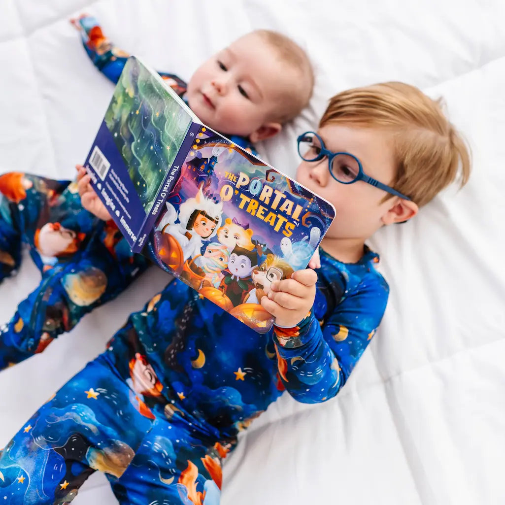 Two children in matching blue pajamas holding a book titled 'The Portal of Treats' on a white blanket.
