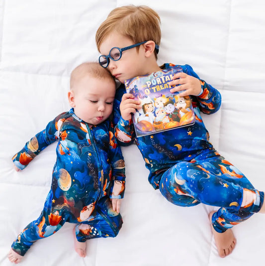 Two children in matching space-themed pajamas holding a book on a white bed.