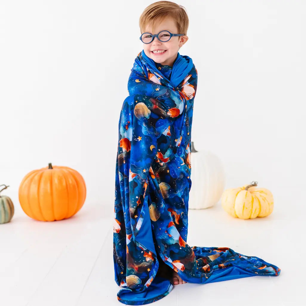 Child wrapped in a blue blanket with space-themed design, surrounded by pumpkins on a white background