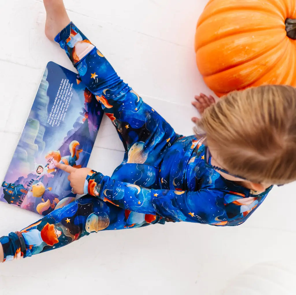Child wearing blue leggings with orange and yellow patterns, sitting next to a pumpkin on a white floor.