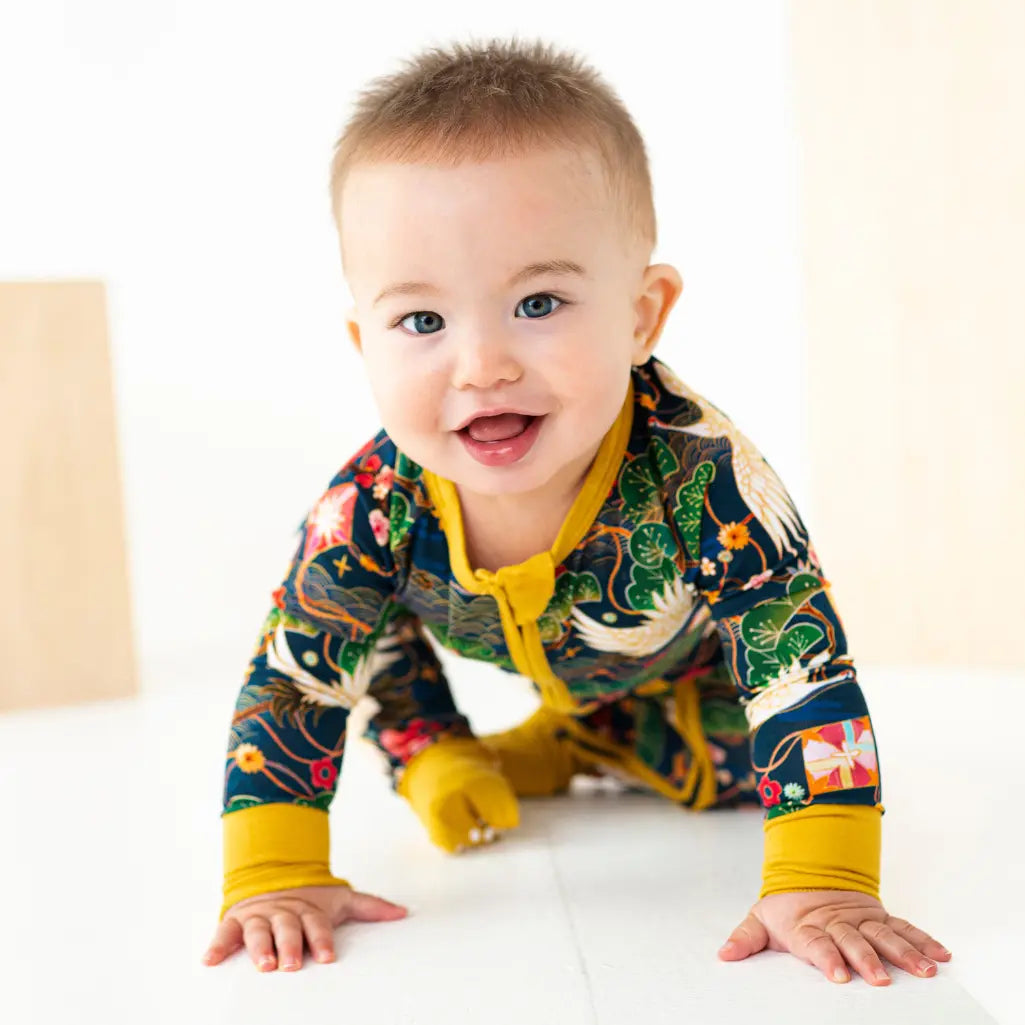 Baby crawling and smiling, wearing a dark floral print romper with yellow cuffs on a white background