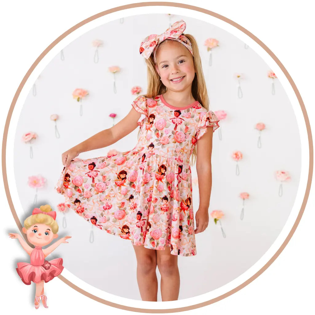 A young girl wearing a pink and red floral print dress with a matching headband, standing in front of a white wall decorated with small pink flowers. The image is framed in a circular border with a cartoon illustration of a girl in a pink dress in the bottom-left corner.