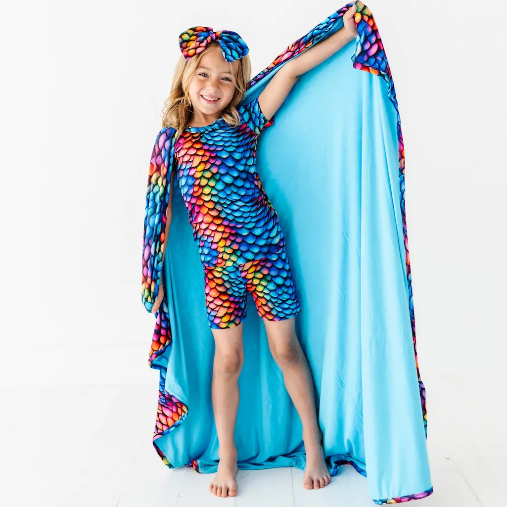 A child holding a blanket with a vibrant scale pattern in rainbow colors, with a matching headband.