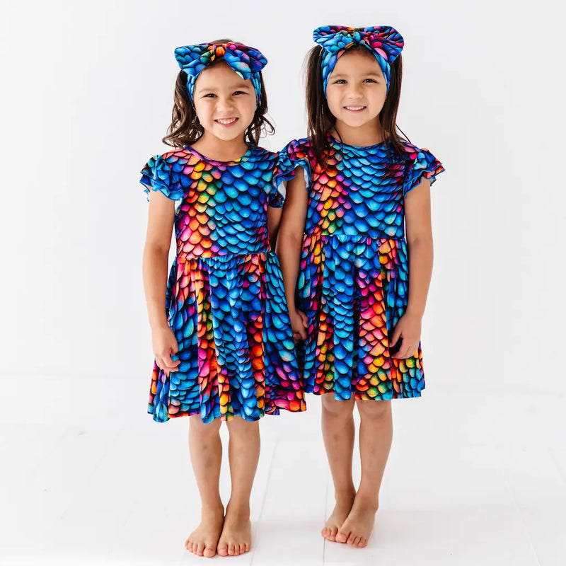 Two children wearing colorful dragon scale dresses and headbands on a white background
