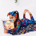 A child wearing a colorful dragon scale-patterned headband and matching outfit, lying down and reading a book.