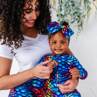 A mother holding a baby, both wearing matching rainbow dragon scale sleepwear, the baby in a matching bow headband.