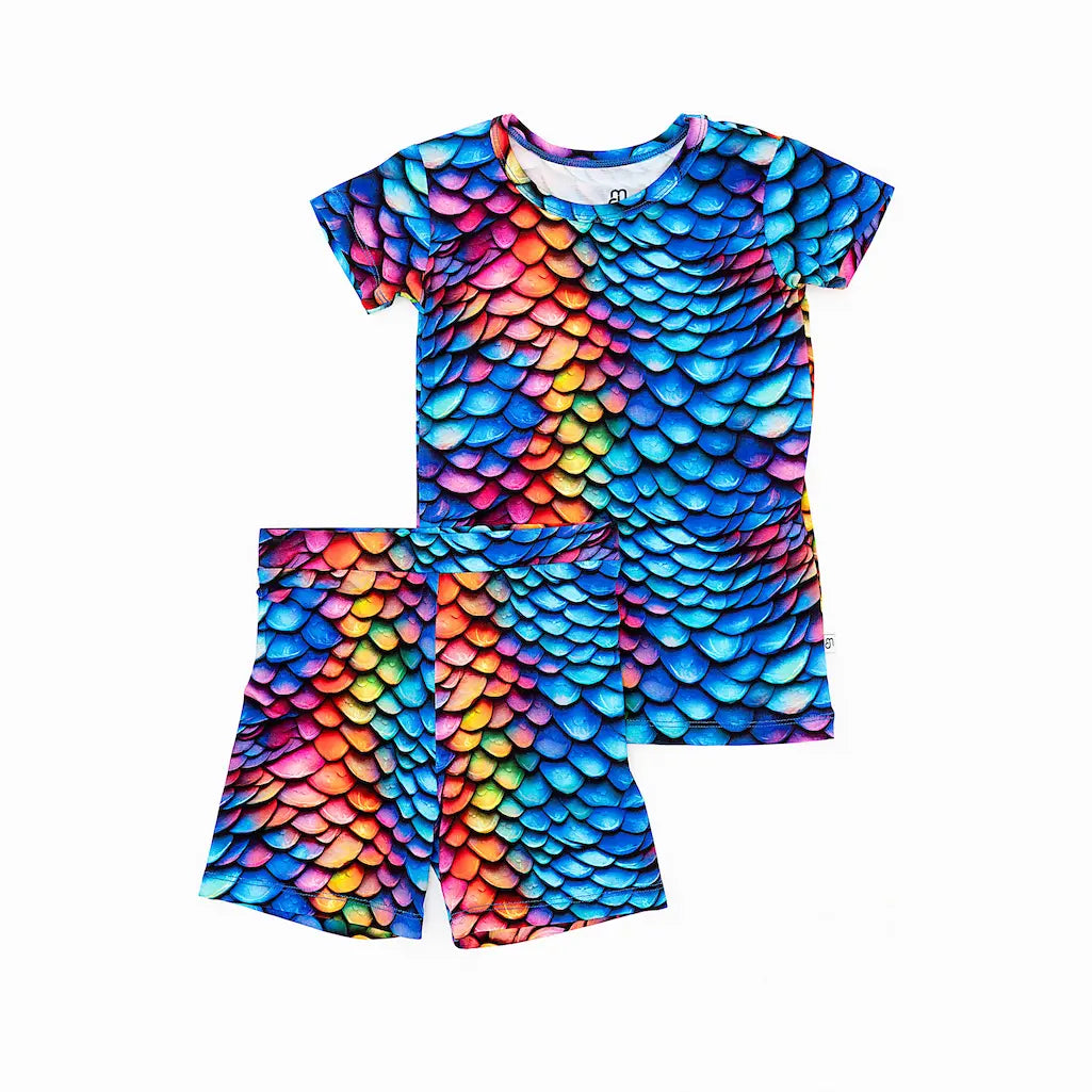 A colorful short pajama set for kids, featuring a scale pattern in rainbow colors, with a top and matching shorts.