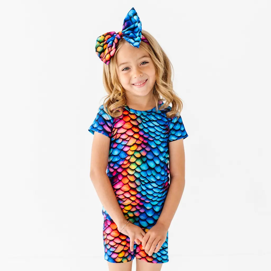 Girl wearing a colorful rainbow scale dress with a matching headband on a white background.