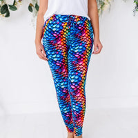 Front view of a woman in a white tee wearing drawstring pajama pants with a rainbow dragon scale pattern.