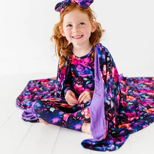 A child sitting on the floor with a multicolored floral blanket around her, smiling.