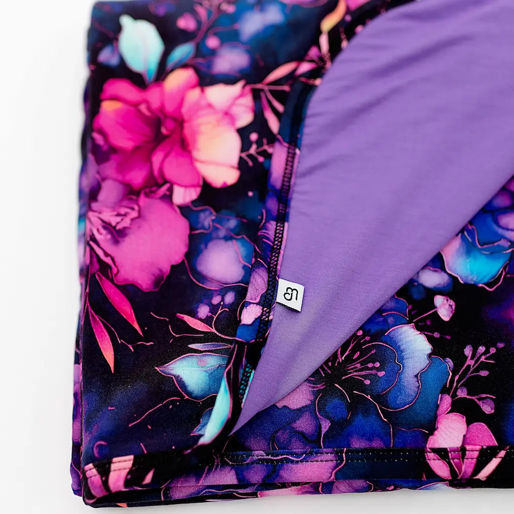 Close-up of a blanket with a whimsical floral pattern in hues of pink, blue, and purple, with a purple solid color on the reverse side.