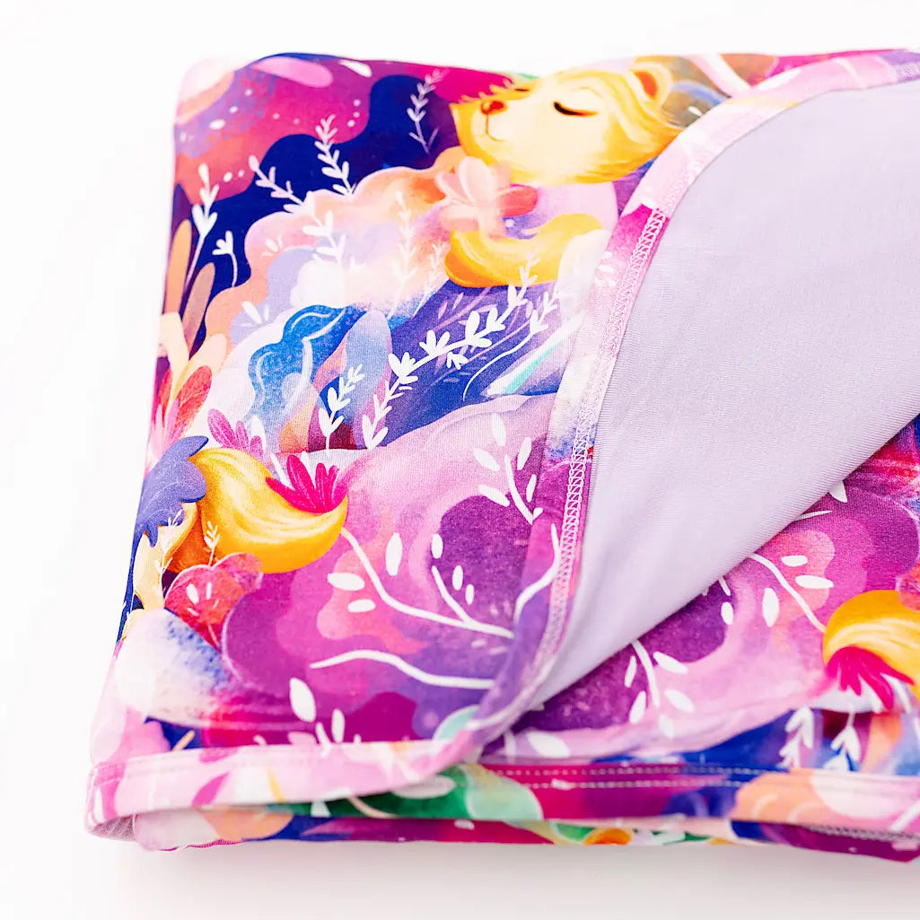Folded plush blanket featuring a colorful floral and unicorn design with a soft lavender underside, displayed on a white background