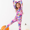 Child wearing a colorful onesie with bunny ears, standing next to pumpkins on a white background