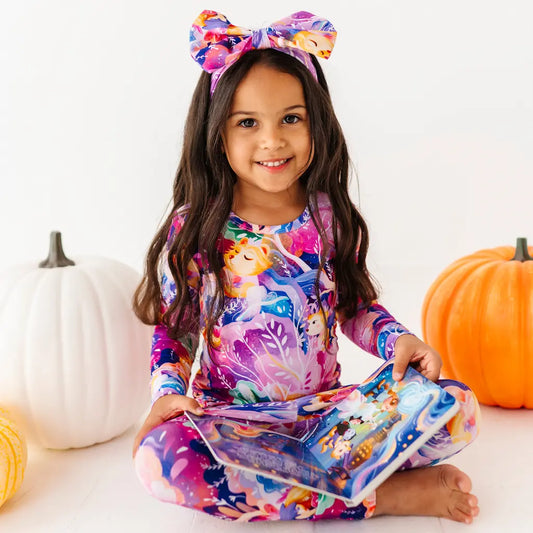 Child wearing a colorful outfit with animal prints and a headband, sitting on a white surface with pumpkins in the background.