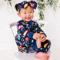 Child wearing a colorful outfit with a floral headband, sitting on a white chair.