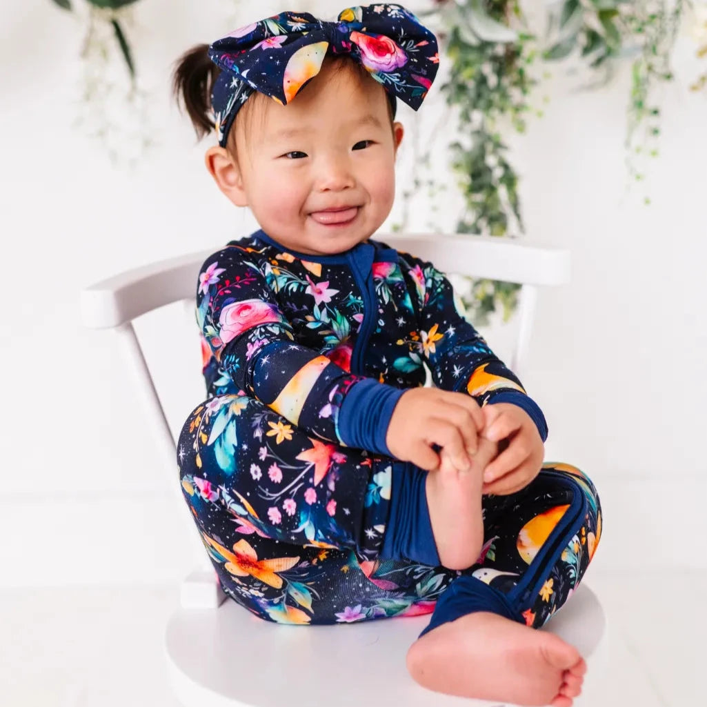 Child wearing a colorful outfit with a floral headband, sitting on a white chair.