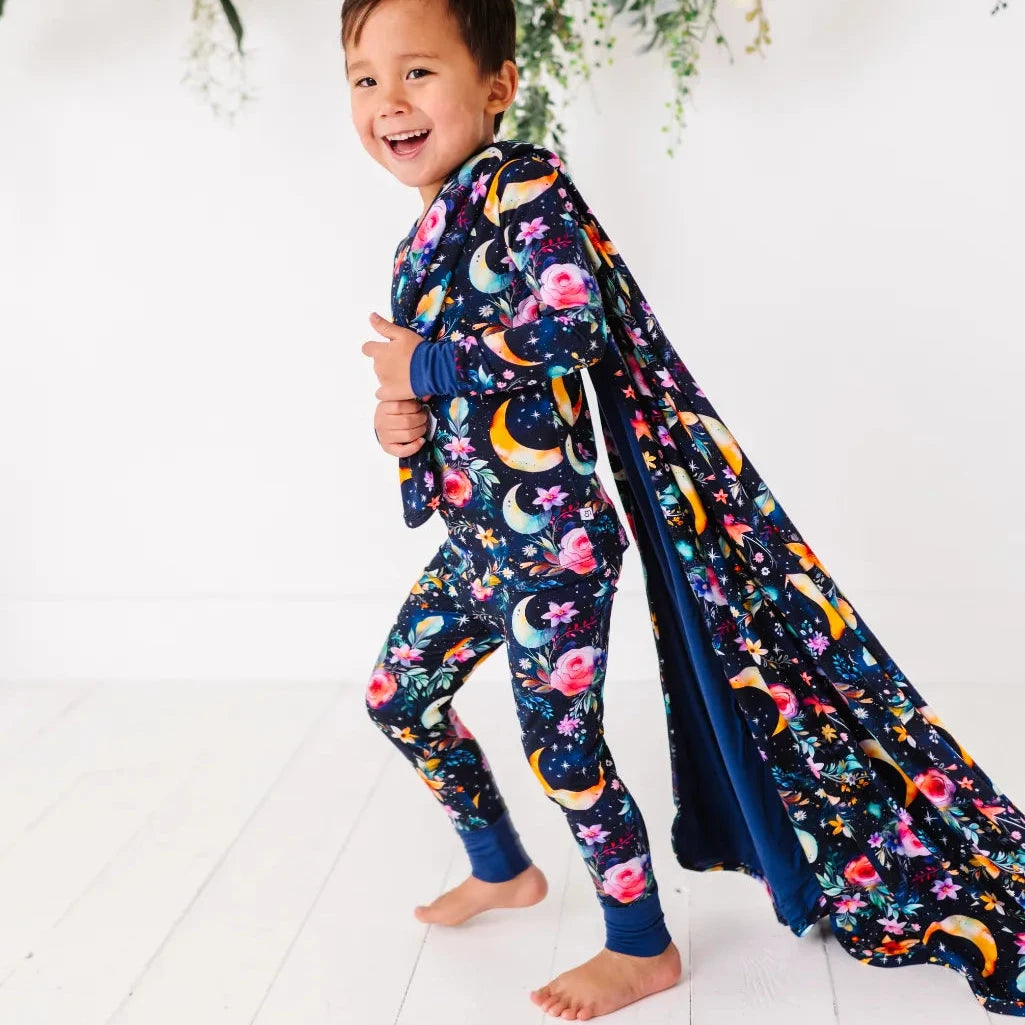 A child standing on a white surface, draped in a navy blue blanket with a floral and animal pattern, smiling and pointing to the side.