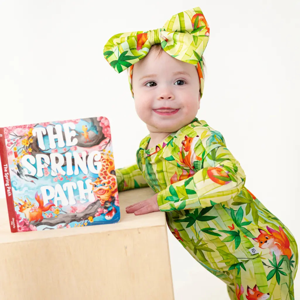 Baby in a green and yellow outfit with a book titled 'The Spring Patch' on a white background
