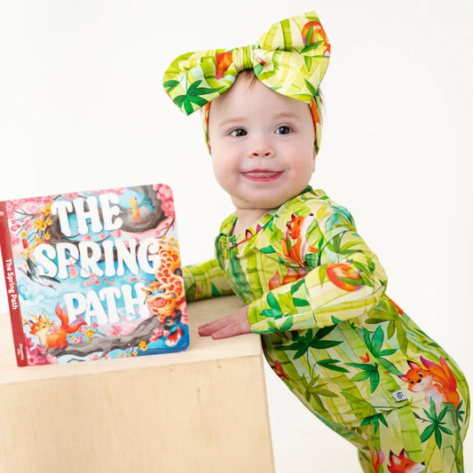 Baby in a green and yellow outfit with a book titled 'The Spring Patch' on a white background