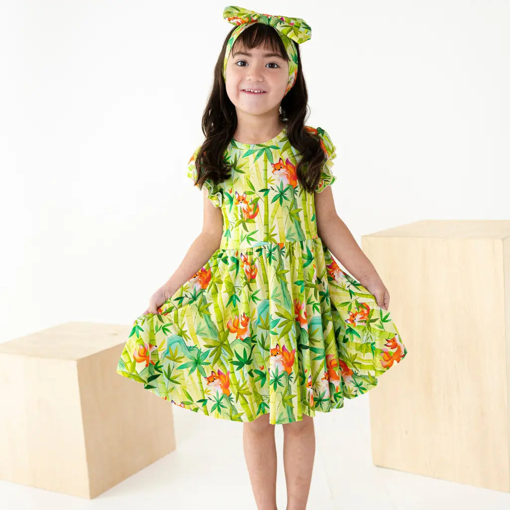 Young girl wearing a green floral dress with a matching headband, standing on a white background.