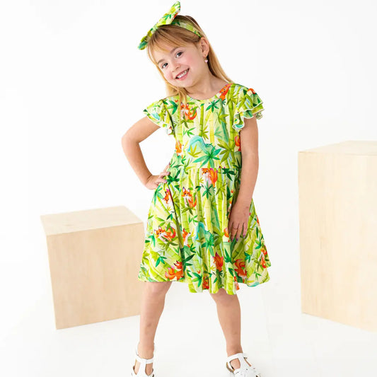 Child wearing a green tropical print dress with ruffled sleeves and a matching headband, standing with hands on hips