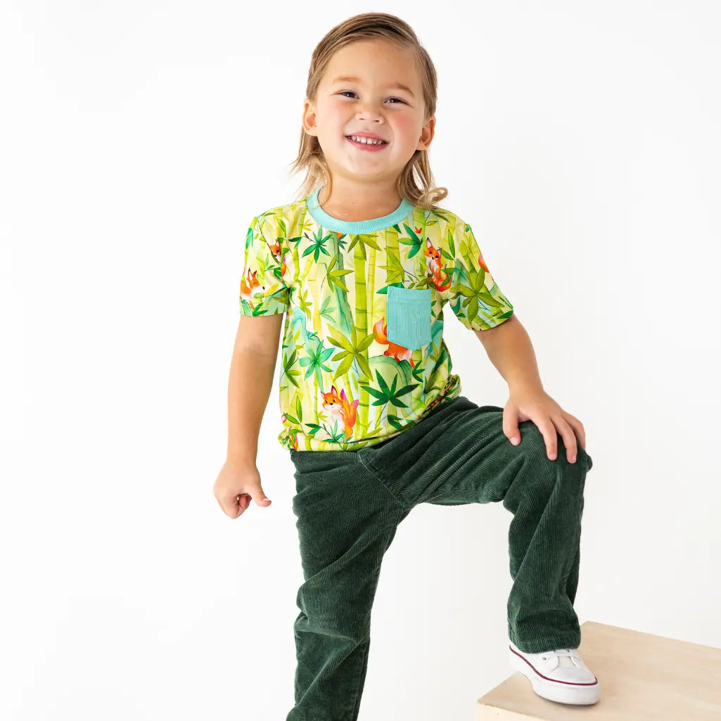 Child wearing a colorful shirt with a dinosaur pattern and green pants on a white background
