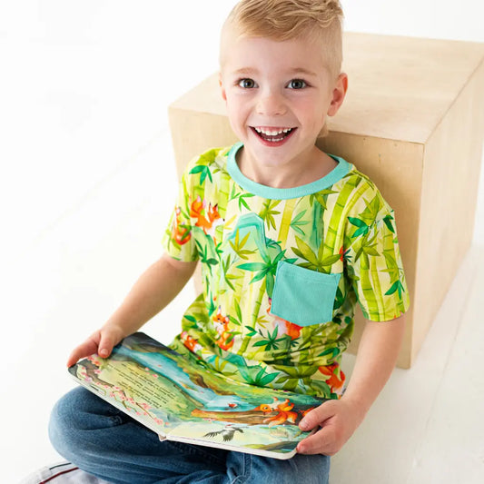 Child wearing a colorful shirt with a nature pattern, sitting on a chair and holding a book.