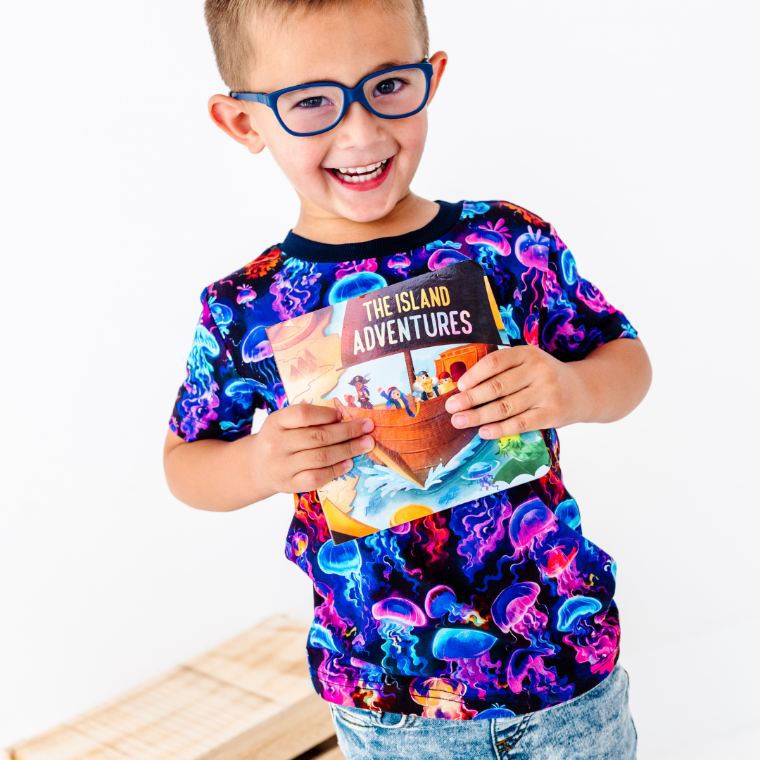 Child wearing a colorful shirt with jellyfish design, holding a book titled 'The Island Adventures'.