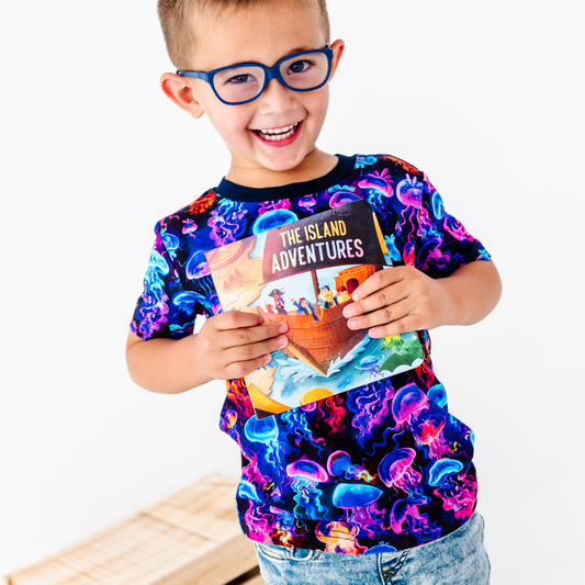 Child wearing a colorful shirt with jellyfish design, holding a book titled 'The Island Adventures'.